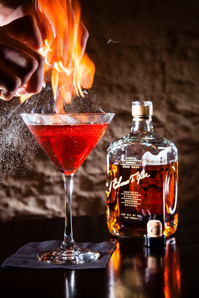 Red cocktail with a burst of flame beside a bottle on a dark bar